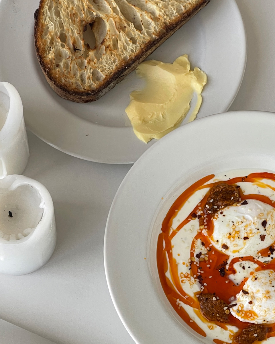 a bowl of turkish eggs in yoghurt with chili oil and a slice of sourdough toast and butter